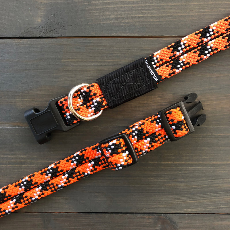 A close-up image of two Wildcat Collars, showcasing their durable orange, black, and white patterns. These collars feature plastic buckles and a metal D-ring for leash attachment, crafted from sturdy materials reminiscent of rock climbing rope. The collars are displayed on a dark wooden surface.
